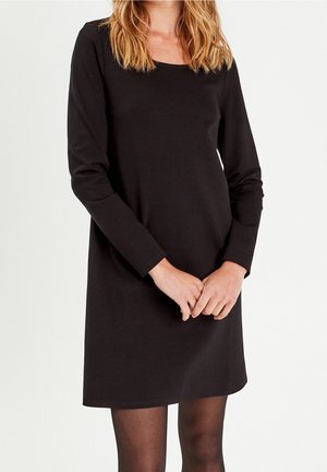 Femme portant une robe noire unie à manches longues et des collants sombres transparents, debout les mains jointes devant, sur un fond blanc.