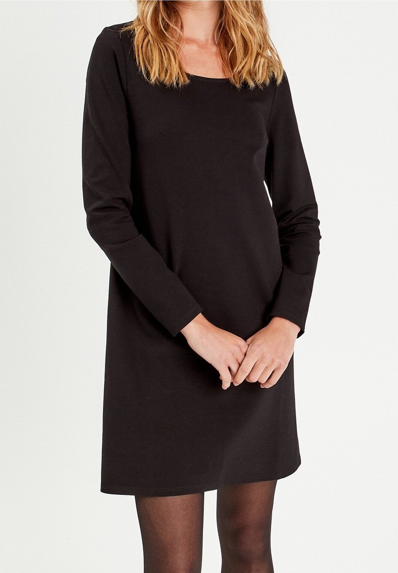 Femme portant une robe noire unie à manches longues et des collants sombres transparents, debout les mains jointes devant, sur un fond blanc.