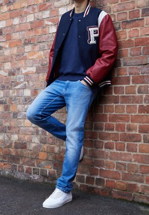 Chaqueta universitaria en azul marino y rojo con un parche de letras, combinada con un suéter azul marino, jeans de un azul claro y zapatillas blancas, posando frente a una pared de ladrillos.