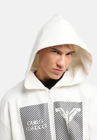 Witte hoodie met een ritssluiting aan de voorkant, zwart-wit grafisch patroon en een capuchon. Het materiaal lijkt zacht en heeft een gestructureerde uitstraling.