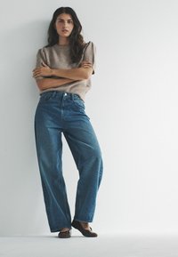 Kobieta stoi ze skrzyżowanymi ramionami, ubrana w beżowy, krótki sweter z krótkim rękawem, niebieskie szerokie dżinsy i ciemne płaskie buty na tle białego, gładkiego tła.