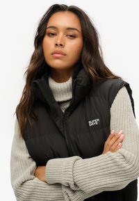 Czarna kamizelka puchowa z wysokim kołnierzem i kieszeniami po bokach. Posiada zamek błyskawiczny z przodu oraz małe logo "Superdry". Nosi się ją na szarej dzianinowej bluzie.