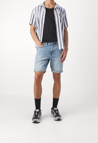 Hombre vistiendo pantalones cortos de mezclilla azul claros remangados, camiseta negra, camisa de manga corta a rayas abierta, calcetines negros y zapatillas negras, blancas y rojas.