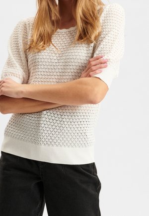 Kvinde med korslagte arme iført en hvid tekstureret kortærmet sweater og sorte bukser, stående mod en ensfarvet lys baggrund.