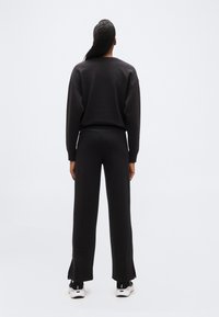 Άτομο που στέκεται γυρισμένο από πίσω, φορώντας μ αύρο φούτερ, μαύρο παντελόνι με φαρδιά μπατζάκια και ασπρόμαυρα αθλητικά παπούτσια σε απλό λευκό φόντο.