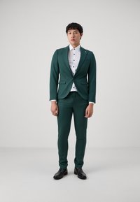 Costume vert avec une veste à un seul bouton et un pantalon assorti, associé à une chemise blanche avec des boutons noirs, complété par des chaussures noires.