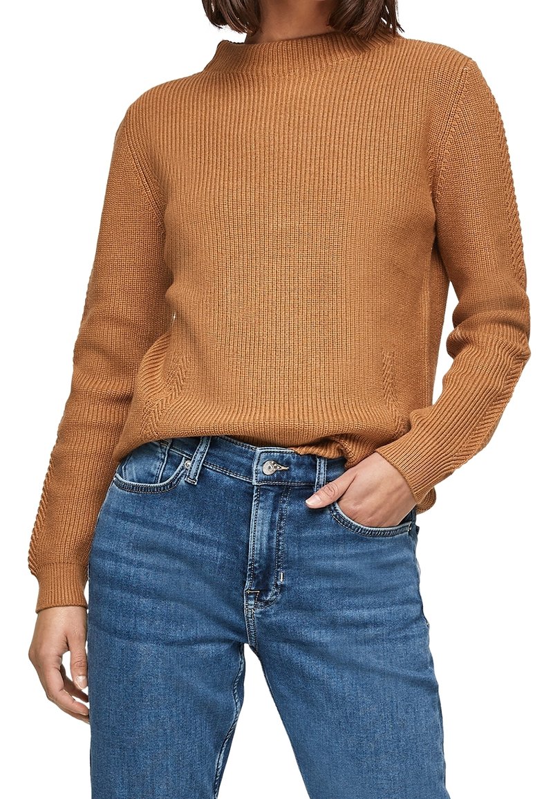 Femme portant un pull en maille marron avec une texture côtelée et un jean en denim bleu, une main dans la poche du jean, sur fond blanc.