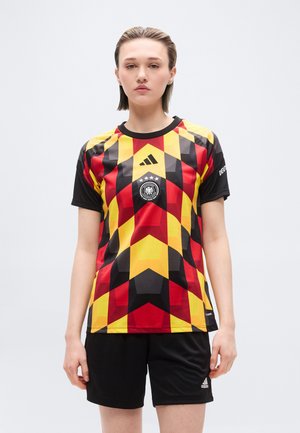Jeune personne aux cheveux courts portant un maillot de football allemand à motifs géométriques rouges, jaunes et noirs, ainsi qu'un short Adidas noir.