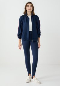 Giacca zip nera con colletto arrotondato, polsini a coste; abbinata a leggings coordinati e scarpe da ginnastica bianche. Texture in maglia liscia in tutto.