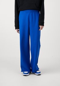 Siniset leveälahkeiset housut, jotka on valmistettu pehmeästä kankaasta ja joissa on joustava vyötärö. Yhdistettynä valkoisiin ja sinisiin tennareihin, mustan hupparin kanssa.