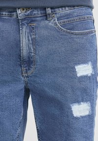 Detalle en primer plano de unos vaqueros de denim azul con detalles desgastados y dos parches rectangulares desgastados en el muslo derecho.
