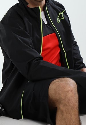 Homme portant une veste noire à bordure néon, une chemise rouge et noire, et un short noir, assis avec un genou visible sur un fond uni.