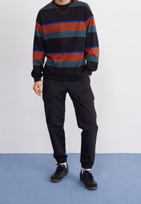 Černá mikina s dlouhým rukávem s červenými, zelenými a modrými vodorovnými pruhy, doplněná černými kalhotami typu cargo a černými teniskami.