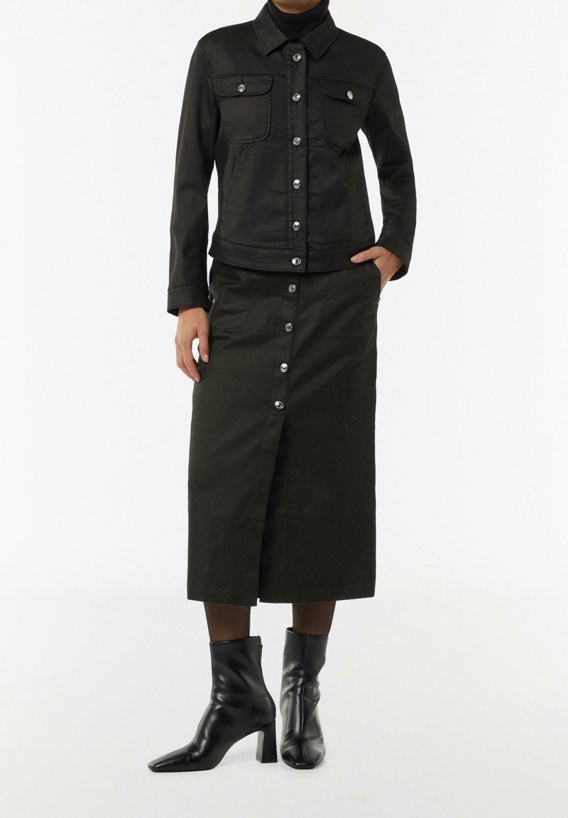 Femme portant une veste noire à boutons, une jupe midi assortie avec une fente avant, et des bottines à talons noires sur fond blanc.