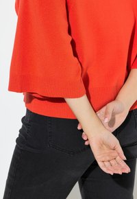 Roter gestrickter Pullover mit geripptem Saum, kombiniert mit schwarzen engen Jeans. Eine Person steht mit hinter dem Rücken verschränkten Händen und zeigt die Passform des Kleidungsstücks.