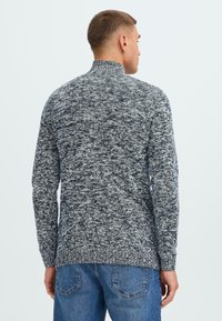 Pull-over en tricot gris et noir moucheté, à col montant et manches longues, associé à un jeans en denim bleu.