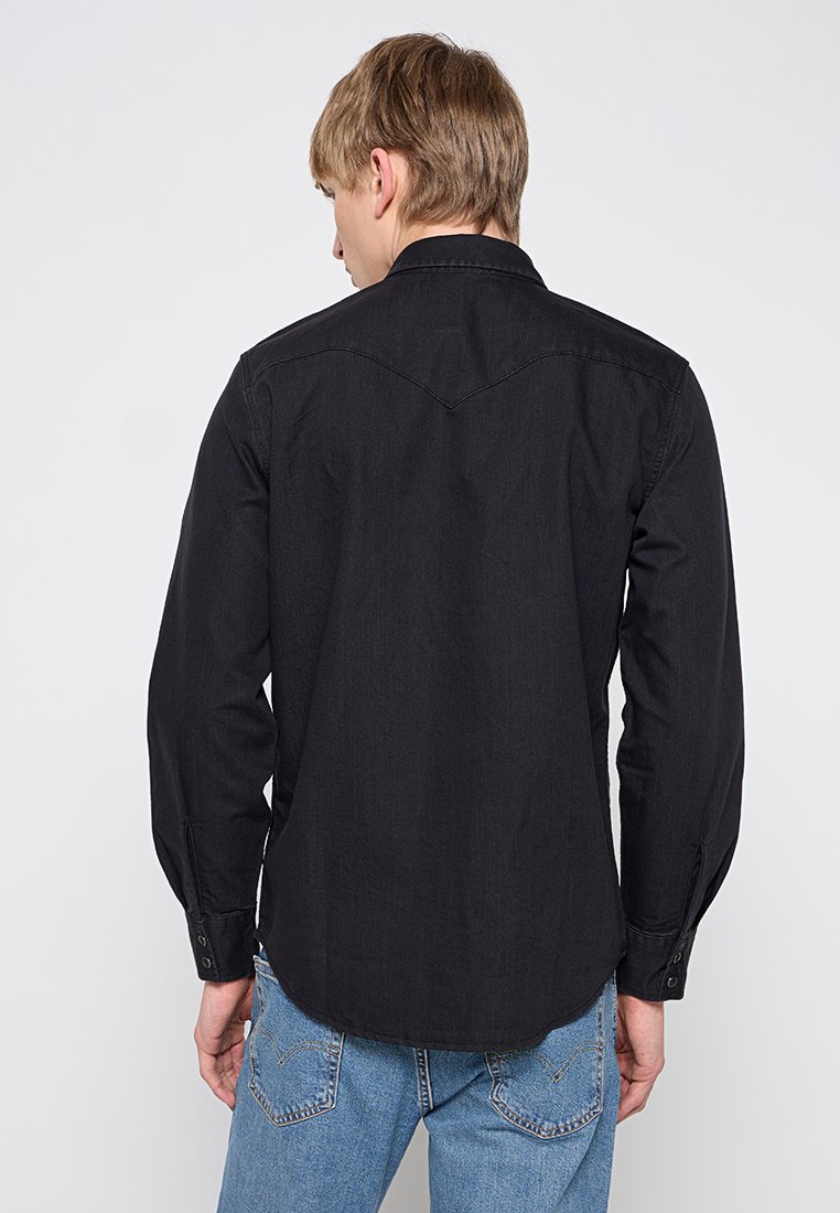 Uomo con capelli corti castano chiaro, vestito con una maglietta nera a maniche lunghe e jeans blu, visto di spalle su uno sfondo neutro.