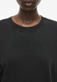 Sudadera negra con cuello redondo, hecha de una tela suave. Presenta un logo tonal sutil en la parte frontal y detalles de costura visibles.
