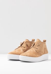 Bottines en suédine beige clair avec une semelle en caoutchouc blanc, présentant des lacets minimalistes et des bords arrondis pour un design épuré.