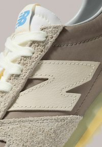 Beiger und brauner Sneaker mit strukturierter Wildlederkappe, glattem Lederlogo, weißen Schnürsenkeln und einer Sohle mit gelben Akzenten.