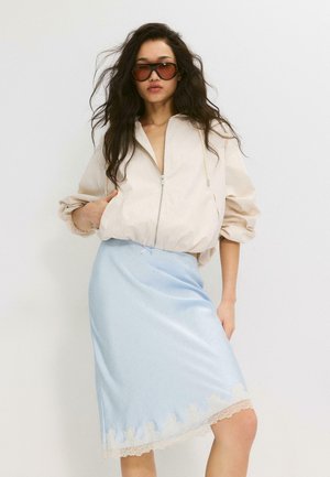 Donna con capelli lunghi e scuri che indossa occhiali da sole marroni oversize, una giacca beige con cerniera e una gonna azzurra con rifinitura in pizzo.