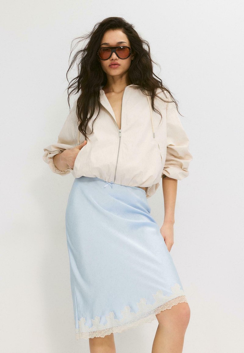 Femme aux longs cheveux foncés portant de grandes lunettes de soleil marron, une veste beige zippée et une jupe bleu clair avec bordure en dentelle.