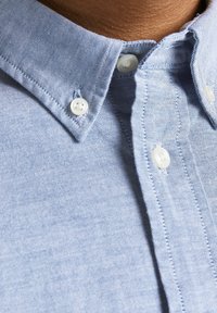 Camisa abotonada de color azul claro con cuello clásico, botones blancos y una tela texturizada. Costuras visibles que acentúan la tapeta.
