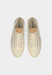 Κρεμ πάνινα παπούτσια Converse με μπεζ δερμάτινη γλώσσα, μεταλλικά ροδέλες και λαστιχένιες μύτες, φωτογραφισμένα από πάνω σε λευκό φόντο.