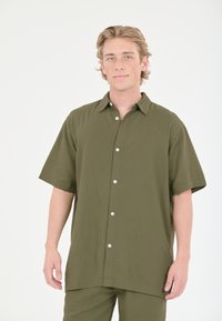 Lyhythihainen, oliivinvihreä nappipaita rentoa leikkausta. Kankaan pinta on sileä, ja siinä on kaulusta sekä valkoisia nappeja.