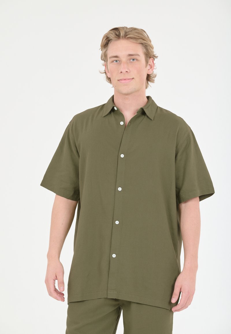 Lyhythihainen, oliivinvihreä nappipaita rentoa leikkausta. Kankaan pinta on sileä, ja siinä on kaulusta sekä valkoisia nappeja.