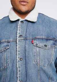 Jeansjacka med ljusblå tvätt, utrustad med en vit sherpa-krage, metallknappar och två framfickor med en liten röd etikett.