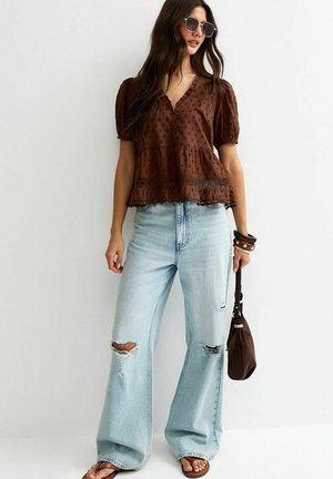 Vrouw draagt een bruine blouse met textuur en pofmouwen, lichtblauwe wijde jeans met scheuren, een zonnebril, een kleine bruine tas en sandalen.