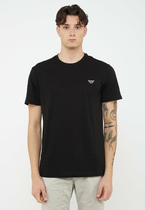 Giovane uomo con capelli corti e ricci, indossa una maglietta nera a maniche corte con un piccolo logo sul petto e pantaloni di colore chiaro.