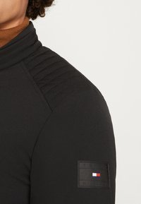 Zwarte jas met een hoge kraag, voorzien van ribbel details op de schouder en een logo op het mouw. Gladde textuur.
