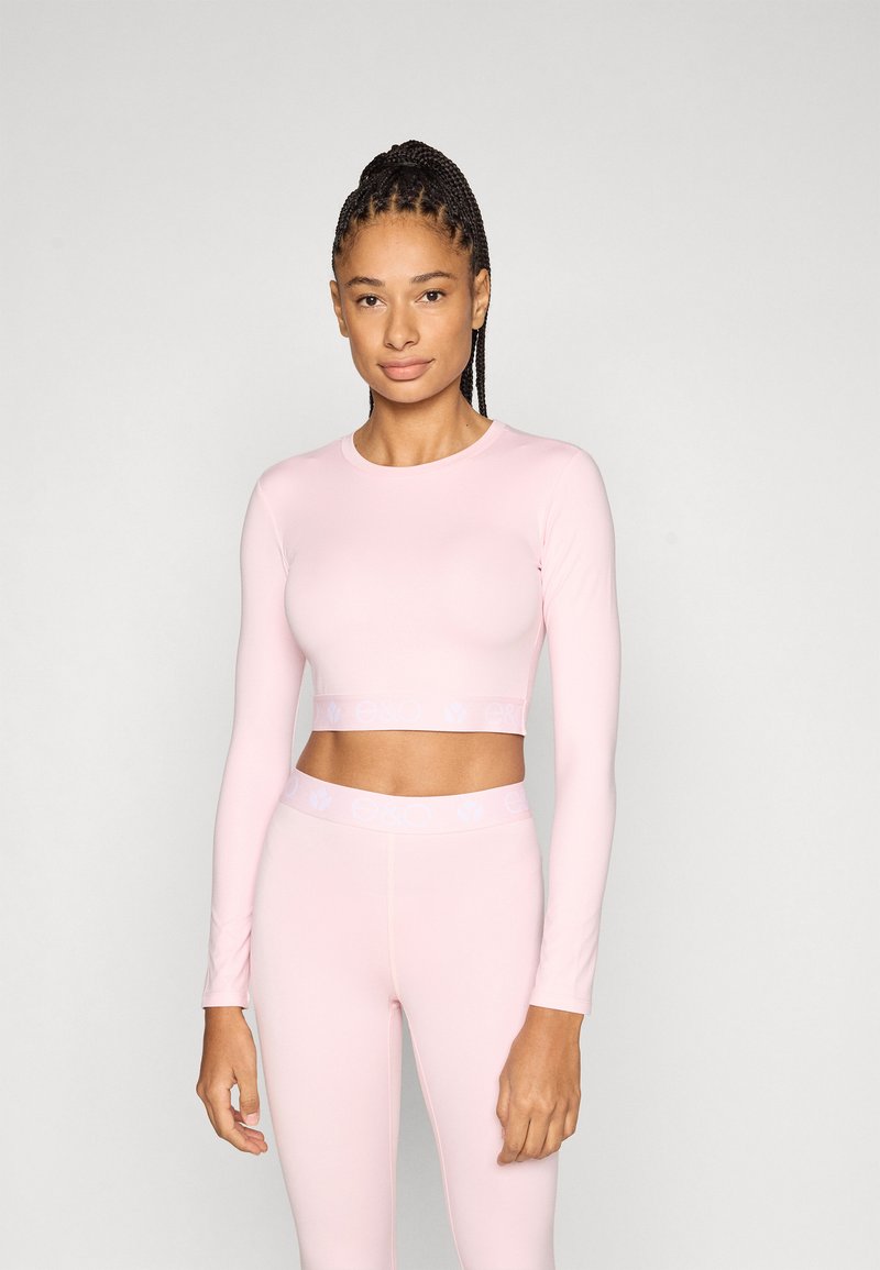 Ροζ ανοιχτό ροζ crop top με μακριά μανίκια, εφαρμοστή σχεδίαση και λωρίδα με λογότυπο. Συνδυασμένο με αντίστοιχα κολάν ψηλόμεσα σε παρόμοιο χρώμα.
