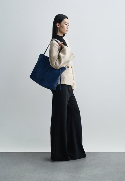 Asian woman wearing beige cardigan and black wide-leg pants, carrying large dark blue tote bag on shoulder, standing against white wall.
