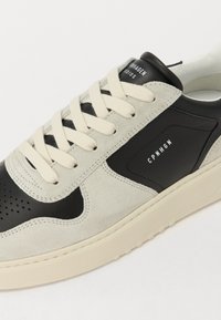 Heled hall nahk ja must nahk sneaker, kreemiliste paeltega, Õhupuuriga sõrmeosa detail ja kummist tallaga. Kõrval on logo.