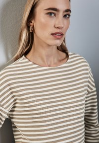 Gestreifter Pullover in Beige und Creme, mit rundem Ausschnitt und strukturierter Stoffqualität. Das Model trägt goldene Creolen.