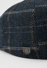 Sötétkék gyapjú sapka kockás mintázattal, bézs vonalakkal. Az oldalán fémes pajzs logó található. Texturált felület.