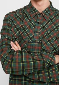 Person klädd i en rutig skjorta i grönt, rött och gult med armarna korsade över bröstet.