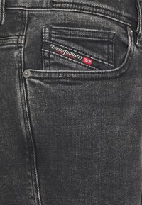 Jeans de mezclilla negra con un bolsillo para reloj que lleva un parche de logo rojo y negro. El herraje plateado acentúa la tela oscura y las costuras.