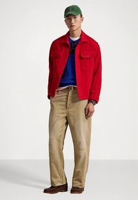 Czerwona bawełniana kurtka, niebieski sweter w warkocze, beżowe spodnie, zielona czapka; posiada kieszenie na guziki, luźny krój i przetarte akcenty na nogawkach spodni.