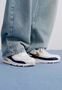 Bijele i tamnoplave tenisice s zlatnim detaljima, koje imaju teksturirani međupod, vezanje na vezice i jastučić s zrakom. Uparene s širokim plavim trapericama.