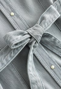 Tissu en denim gris clair avec une ceinture nouée en forme de nœud et deux boutons pression métalliques sur des coutures piquées.