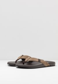 Des tongs brunes avec une sangle texturée et une semelle douce. Les sangles présentent un design croisé avec un accent plus foncé sur le dessus. Semelle légère.