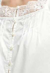 Gros plan d'un haut blanc avec un col en dentelle délicate et une rangée verticale de petits boutons blancs sur le devant.