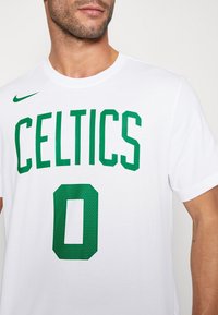 Homme portant un t-shirt blanc avec un texte vert "CELTICS" et le chiffre zéro, avec un logo Nike vert en haut à gauche.