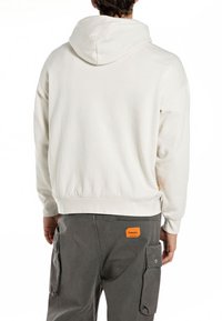 Sudadera blanca con capucha y un corte relajado, con cintura de canalé y etiqueta de logo en la parte trasera. Combinada con pantalones cargo grises que tienen bolsillos laterales.