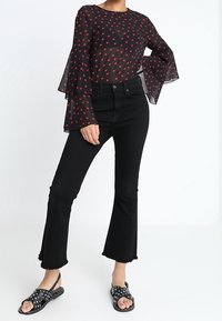 Blouse noire en tissu transparent avec motifs de cœurs rouges, manches évasées ; associée à un jean noir taille haute à ourlet effiloché et à des sandales noires à pois.