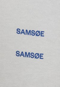 Tessuto bianco con una texture liscia che presenta due linee di testo ricamato blu con la scritta "SAMSØE".